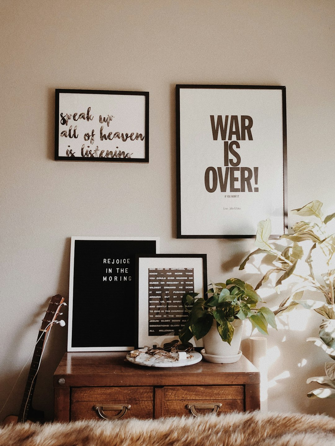 A wall with three framed posters on it, one black and white poster of the text “war is over! talking about heaven”, two color posters in different sizes, a painting in the style of natural light, minimalist, plants, warm colors, a wood table top with small decorations, a guitar leaning against the wall, white walls.