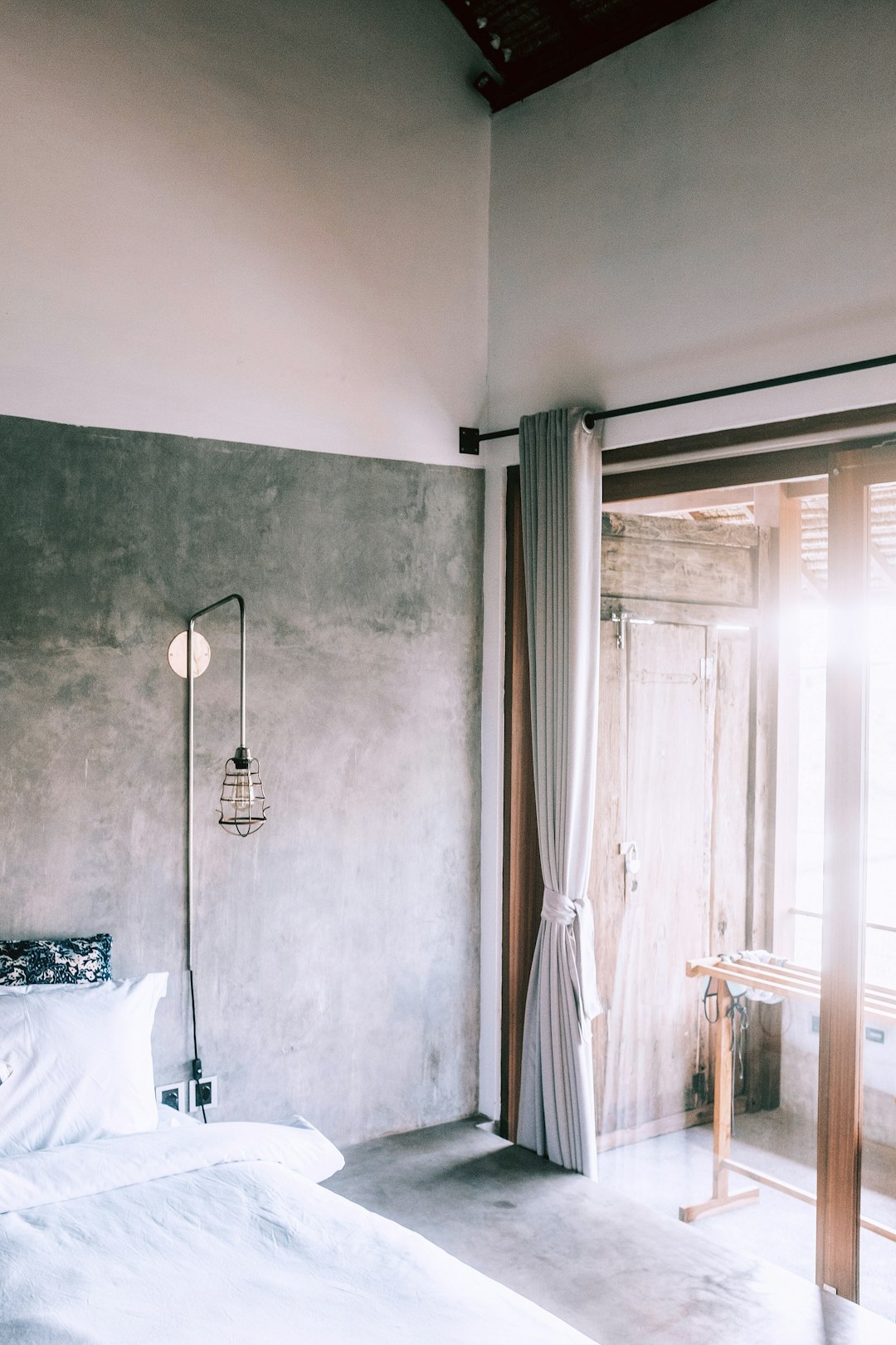 A photo of the bedroom, featuring concrete walls and white curtains on one side. The light grey cement floor, while sunlight shines through an open door onto the bed with white sheets. A hanging lamp hangs from above. There is also a window in front that reveals another room behind it. This photo was taken with a Canon EOS using Kodak Gold film. It has an aesthetic style reminiscent of minimalism in the style of Canon EOS.