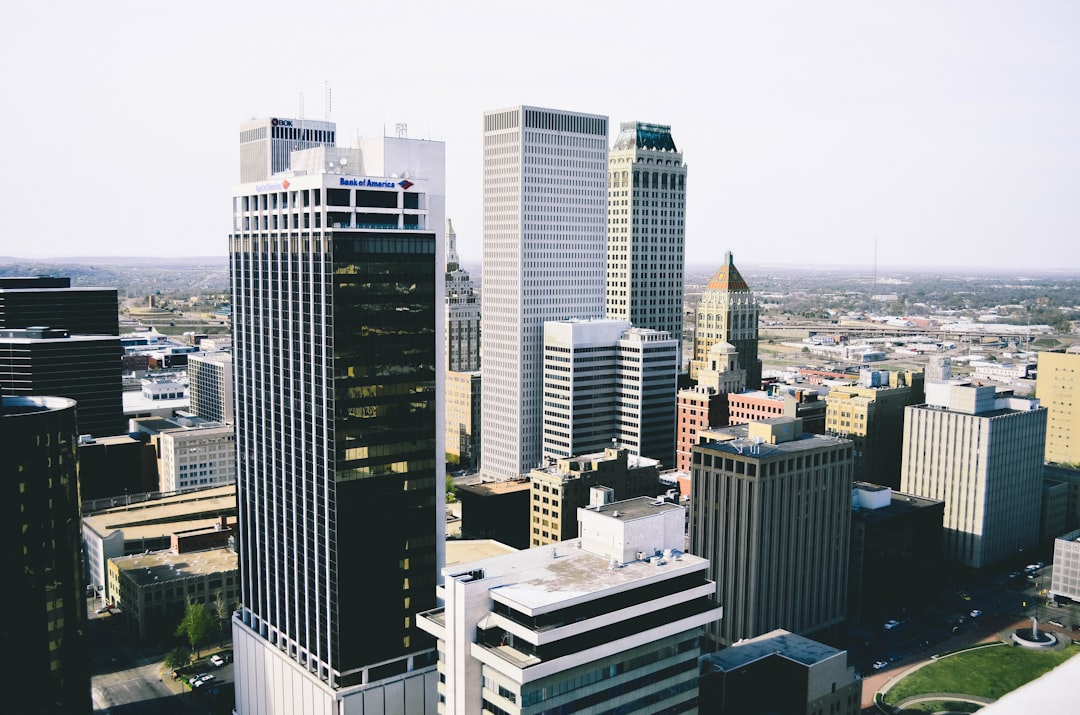 A photo of the cityscape of okc, with tall buildings and skyscrapers. In the center is “okurgeon” written on one building’s facade. The view shows downtown modern architecture, including black glass towers and white walls, contrasting against green grassy hills in the distance. It captures an aerial perspective from above looking down at this urban landscape. Taken during daylight with natural light, shot in the style of Sony Alpha A7 III camera using Kodak Gold film.