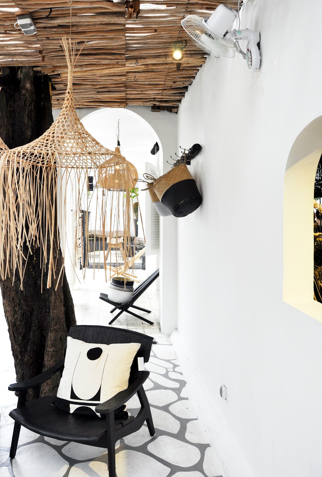 A photo of the entrance to an interior design studio in Oia, Crete with white walls and black accents. The ceiling is made from natural materials such as wood or wicker hanging lights above it. A tree trunk has been cut down at one end and decorated with woven fabric for seating. In front there’s a modern chair with a pillow on top. There’s also a small table next to that with two chairs around it. On each wall hangs a basket containing various items like pillows and bags.