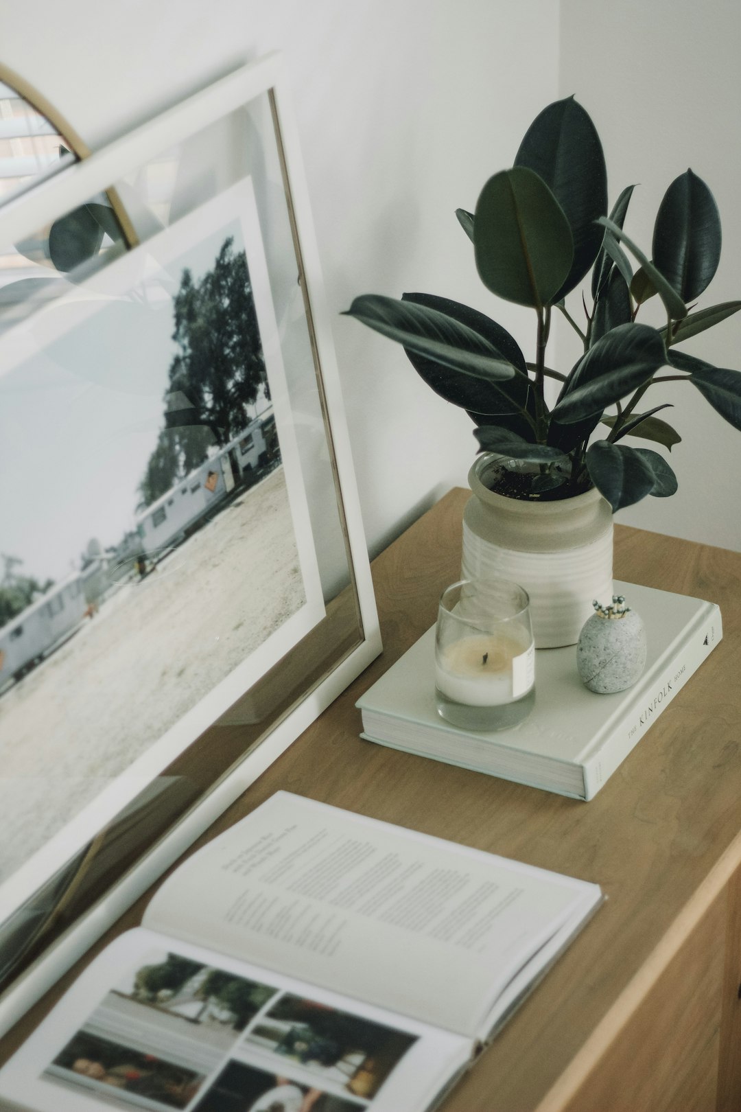 A close up photo of a book, magazine and framed photograph on the desk with white lighting, a small green plant in a vase, a scented candle, in the style of modern home decor, aesthetic, minimalist, neutral colours.