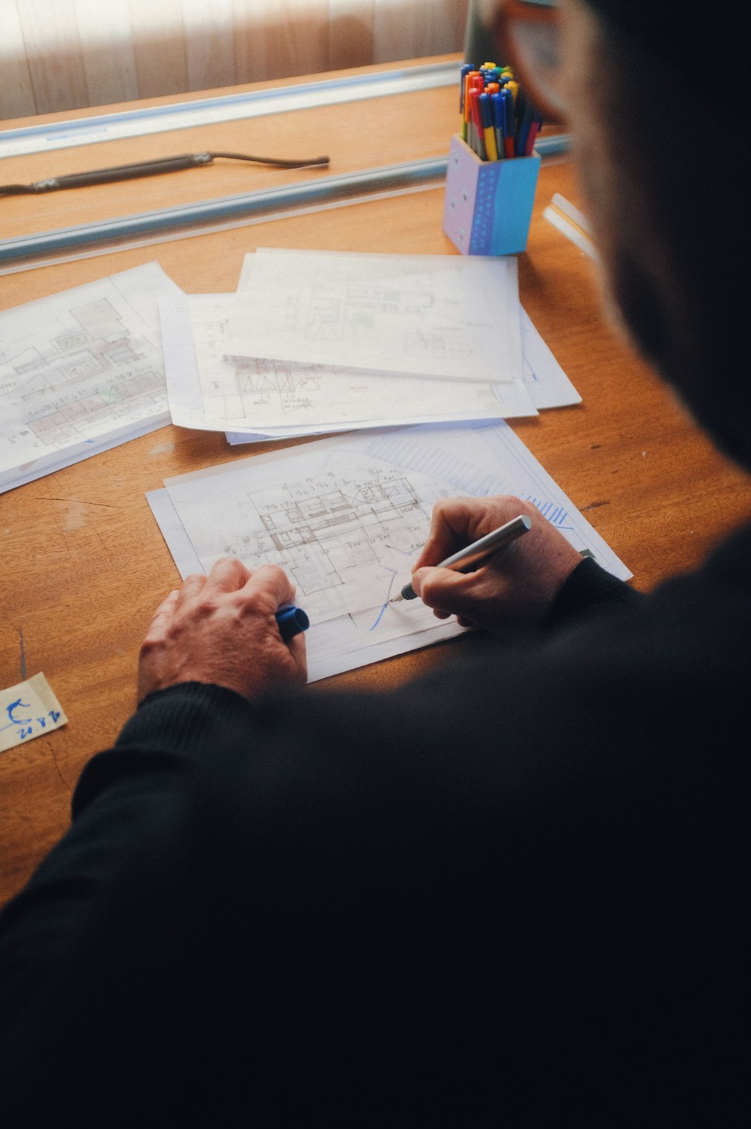 A close-up photo of an architect drawing at his desk, with paper and pens around him. There are some blueprints in the background, shot from above with a Canon EOS R5 at F2, ISO40 3d.