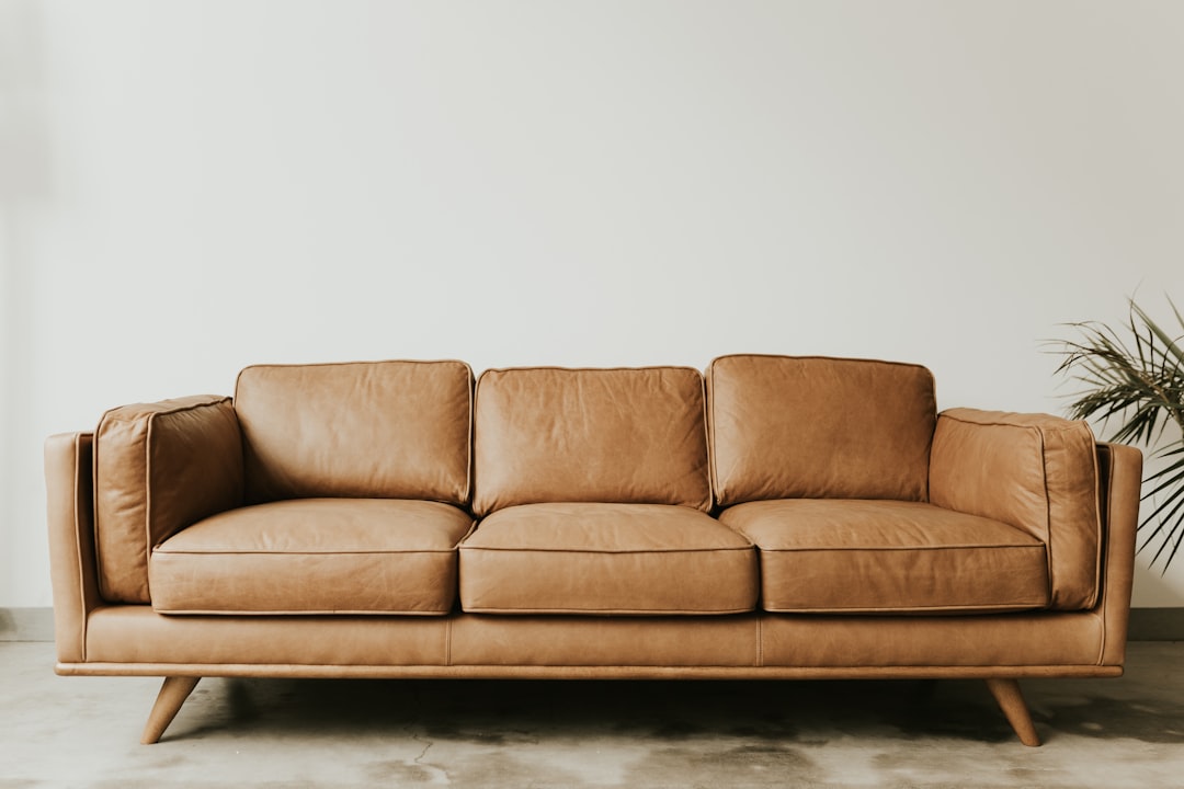 Photograph of an elegant three seater leather sofa in a light brown color, placed against a white wall with minimalist decor. The focus is on the soft texture and warm tones of the couch, creating a cozy atmosphere for seating and relaxation in the style of minimalism.