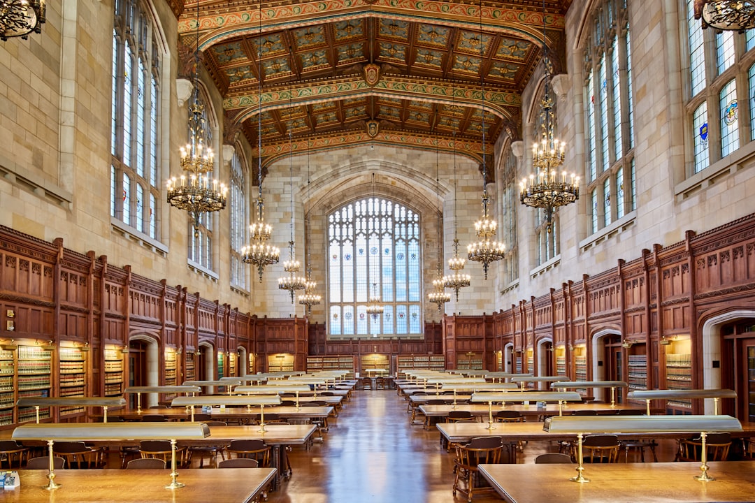 A grand, ornate library hall within the University of Michigan’s had a sidequave ceiling and walls covered in dark wood paneling with many large windows on one side. The space was filled with rows of long tables made from light wooden planks with brass legs that seated multiple chairs each, all lit by chandeliers hanging above them. In front of these tables stood two bookshelves, also made out of thick wooden panels with glass doors at their top to display books. There should be no people present. –ar 128:85