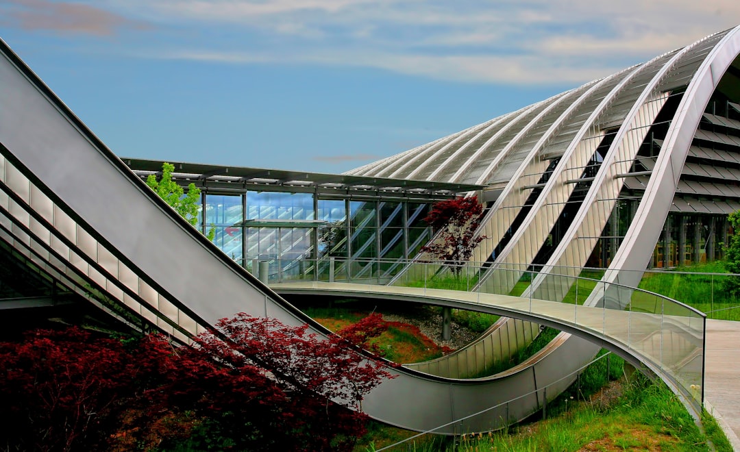 A modern building with an angular roof, glass walls and metal accents. The entrance is through the curved bridge leading to it from another part of the museum complex. There is greenery around, trees in red and purple colour, blue sky in the background. A view from the side on one level showing plants growing inside. It seems like some part of its interior has turned into a botanical garden or park. , architecture photography, architectural visualization in the style of [Iwan Baan](https://goo.gl/search?artist%20Iwan%20Baan).