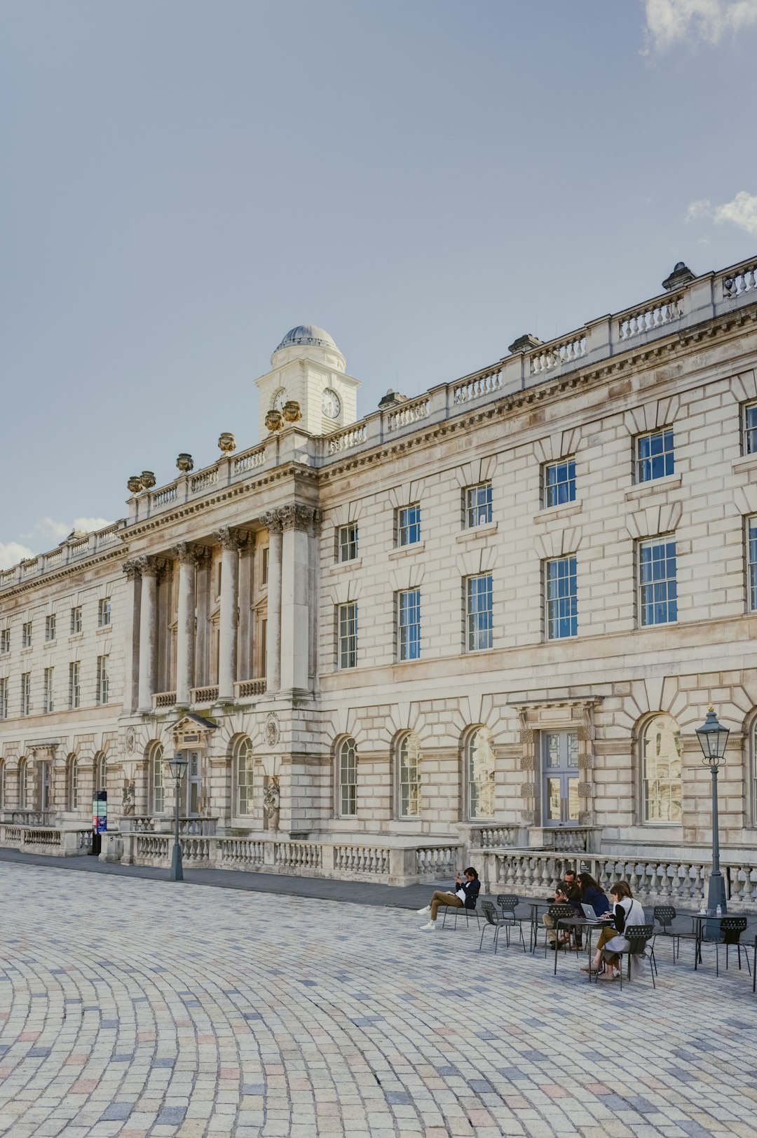 The grand facade of Somerfield Place in London, with its large white stone building and blue sky above. People sit at outdoor tables on the wide cobblestone plaza nearby, while another person walks past the entrance to the main door. The scene is captured from an eyelevel perspective, showcasing both buildings and crowds in the style of high resolution photography.