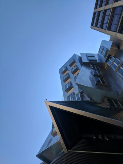 A modern building designed in the style of [Frank Gehry](https://goo.gl/search?artist%20Frank%20Gehry), photographed from the ground looking up at it, against a clear blue sky. The photo was taken with an iPhone and has a slight graininess to it due to low resolution. It captures the unique architecture of it which includes geometric shapes and metal elements on its exterior walls. In the background is another tall building that contrasts with "I3 Newton", adding depth to the scene.