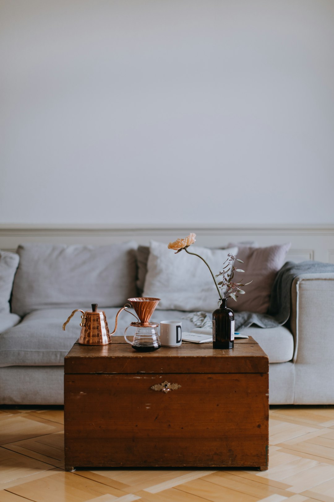 A coffee table in the middle of the living room with an old wooden chest on top. A coffee pot and cup on a side table, with a grey sofa behind. The floor is wooden, in the minimalist style. A low angle shot was taken from above, with a white wall background and natural light. The photography was professional, with boho colors that were simple and modern. –ar 85:128