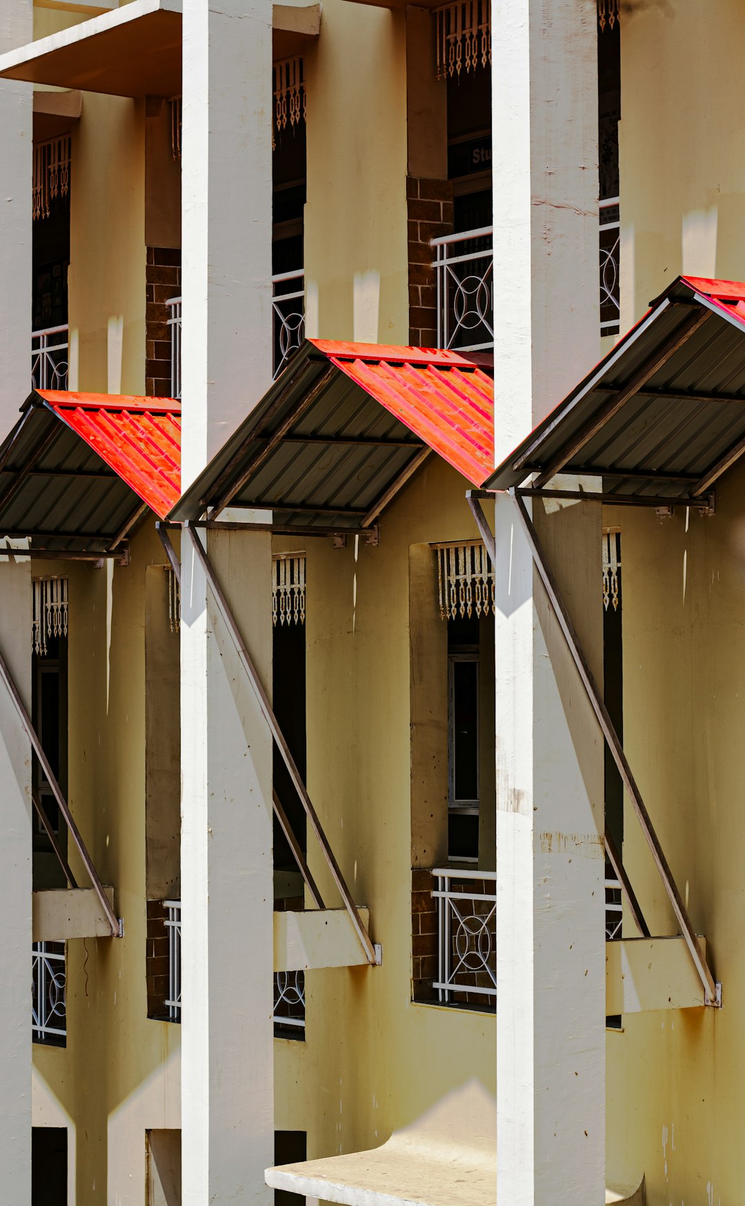 a view of the balconies on an apartment building in kerala with red steel feet and white columns, white walls and yellow floor, the roof is made up by a slanted metal sheet –ar 79:128