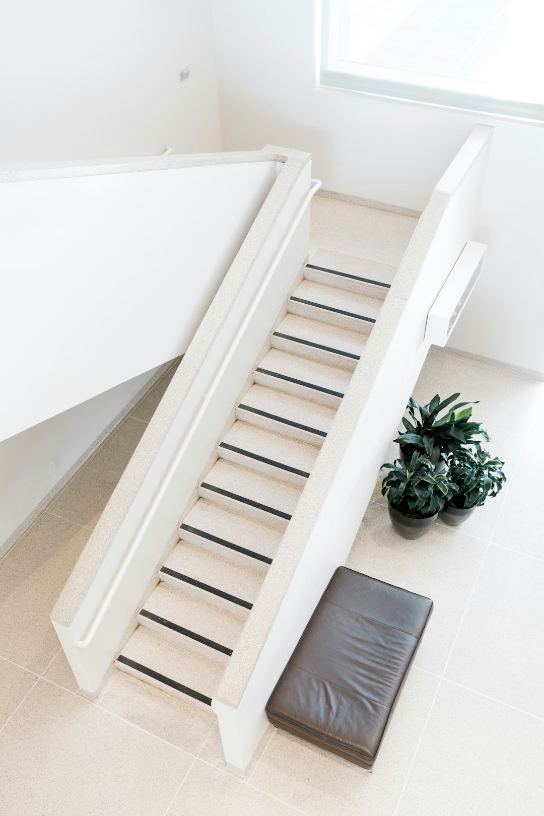 A white staircase with black stripes leads to the bathroom, next to it is an open leather pillow on which one can lay their head and relax. The background features a minimalistic modern interior design in light colors with minimalism, a concrete floor, and plants.