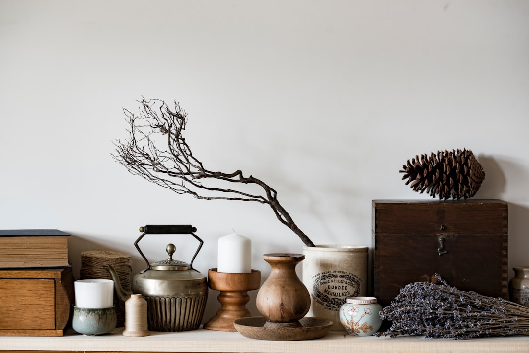 A minimalistic shelf with antique objects, wood and ceramic decorations, a small branch of dried lavender, an old wooden tea kettle, some candles, a chest full of books with a pine cone on top, against a neutral white wall in the background, illuminated by natural light from a window, captured in the style of Canon EOS R5 at F2, ISO400, Shutter speed; 36mm f/8.
