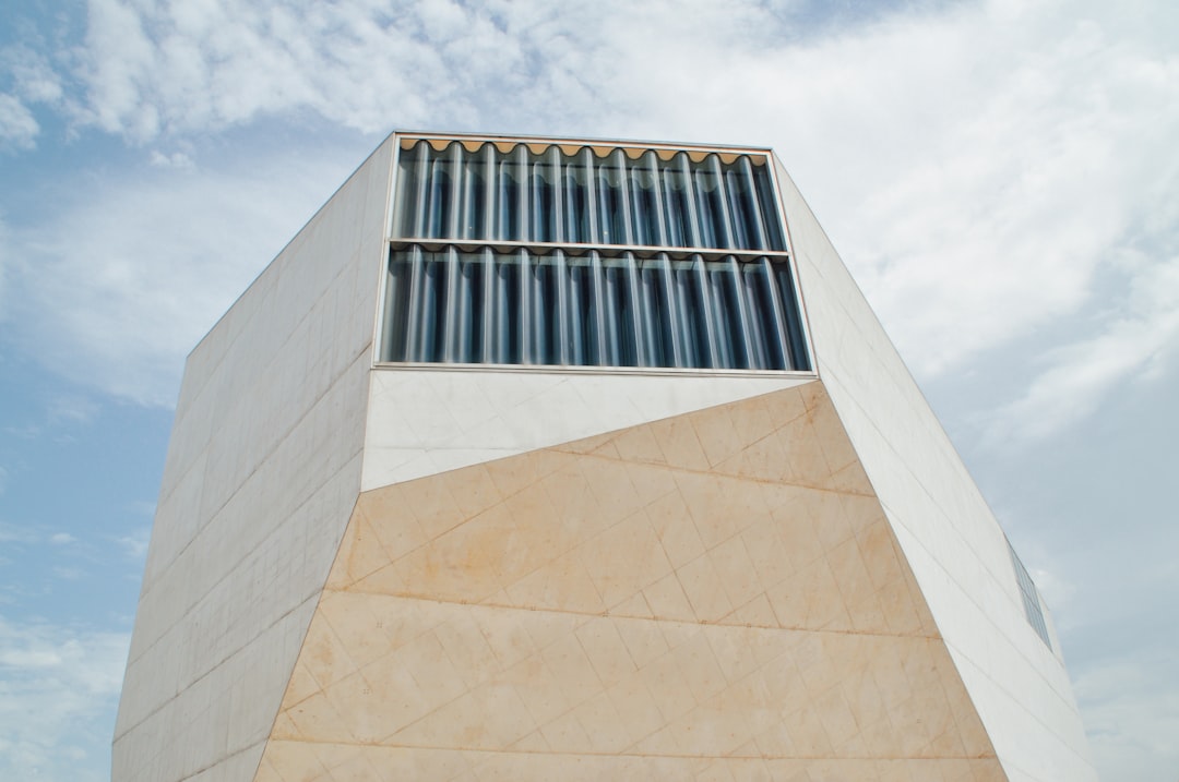 A triangular-shaped building made of light-colored marble, with large vertical windows at the top and an exterior wall that opens up to reveal the interior space. The background is a blue sky with white clouds. Use a Canon EOS camera with a wide-angle lens for a full view of the scene. –ar 128:85