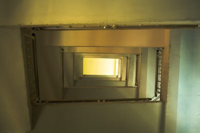 Looking down at the top of an empty staircase, this interior shot shows a soft yellow light coming from above in a square opening. With a straight on view and eye level perspective, no people are present in this architectural photography shot in the style of no particular artist.