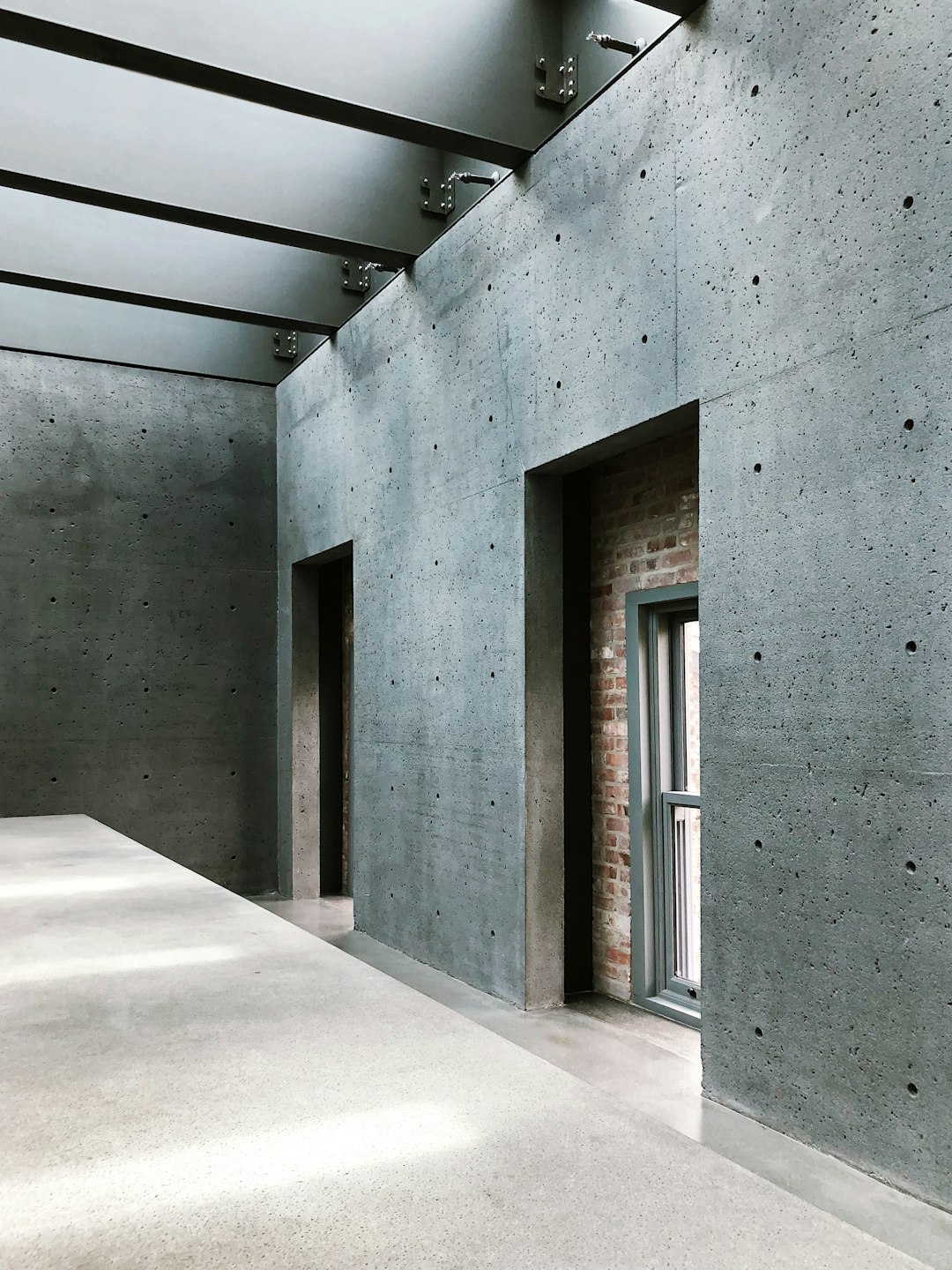 The concrete museum building’s interior view has a tall ceiling with gray walls that have small brick details. A metal door frame is in a long and narrow room with a wide perspective and cool tones under natural light, creating a calm atmosphere.