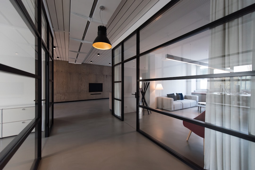 A modern office interior with black metal frame glass doors leading to the living room, concrete walls and white ceiling, large windows, soft lighting, neutral color palette, minimal furniture, clean lines, open space design, architectural photography in the style of an architectural photographer.