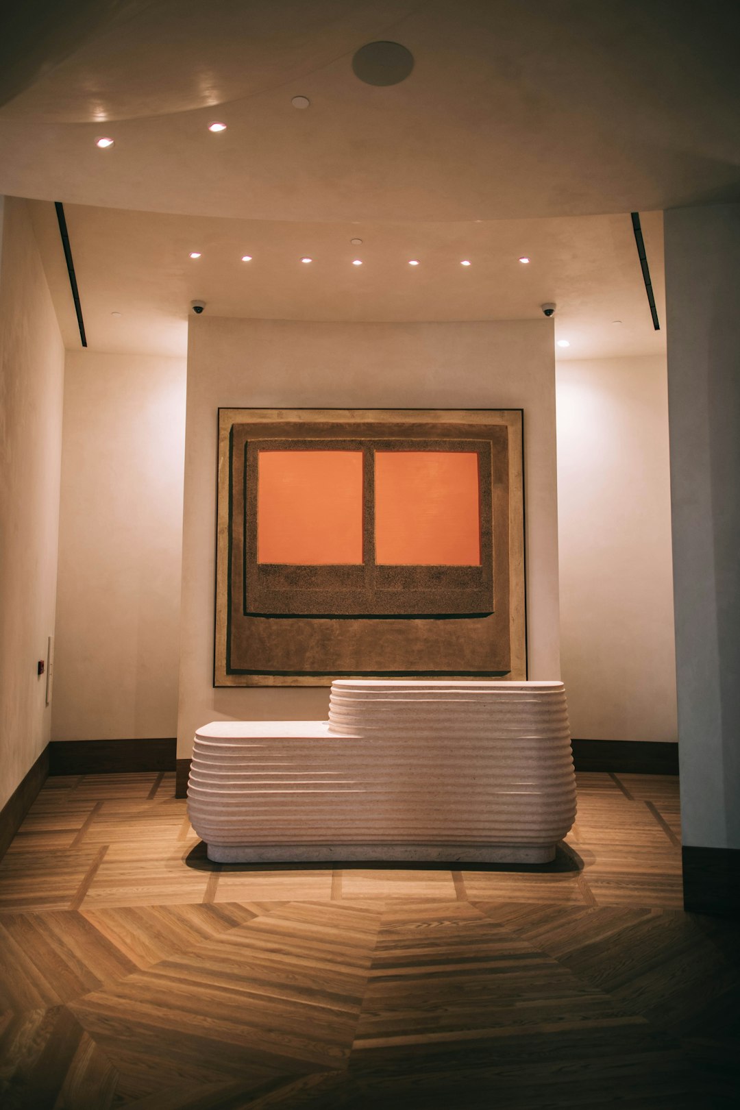 A modern reception desk made of white stone in the center, flanked by two orange paintings with wooden frames, against a wall in an elegant hotel lobby. The room has wood floors and is illuminated by soft lighting. A large window on one side allows natural light to enter. This photo was taken using a Canon EOS R5 camera. –ar 85:128