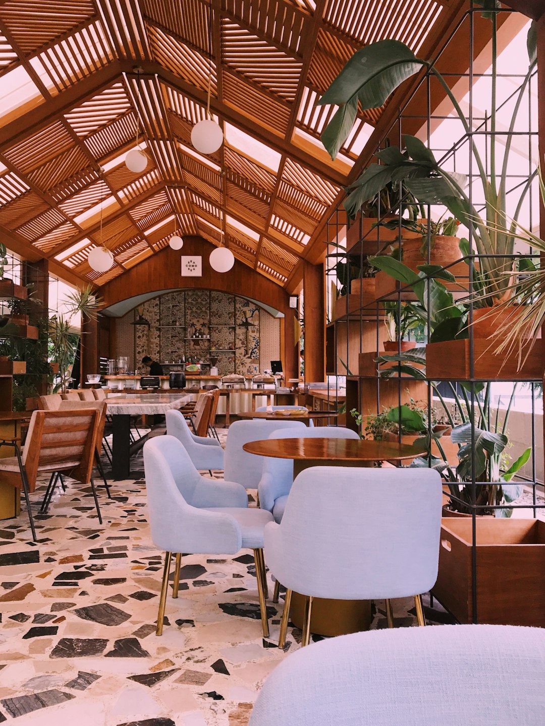 photo of the interior design of a cozy cafe in a biophilic style with wood and terracotta details, white chairs, wooden tables, plants on shelves, blue lightings, a ceiling made from glass roof tiles, a marble floor, sunlight coming through skylights, high ceilings, interior photography. –ar 3:4