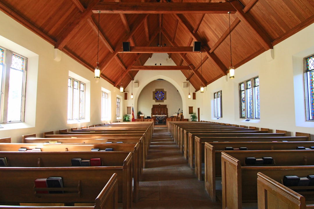 An empty church with pews and a wooden ceiling, looking towards the altar. The photo is taken from behind rows of comfortable benches, providing an expansive view into the interior space. There is natural light streaming in through large windows on either side, creating a warm atmosphere. A small window at one end reveals another room beyond, adding depth to the scene. This photograph captures the serene ambiance within the peaceful chapel setting in the style of a minimalist artist. –ar 128:85