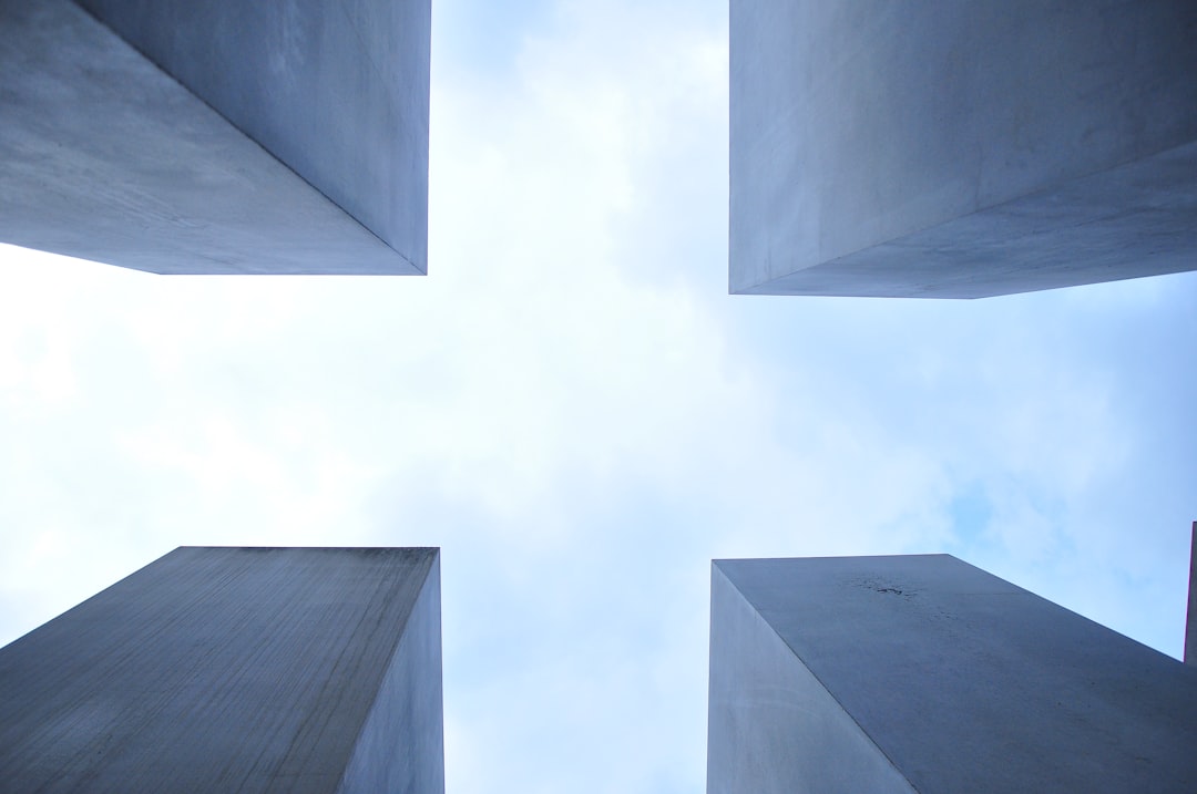 photograph of the outside of each side and bottom corner of four concrete monoliths at varying heights, the sky is blue with white clouds, shot from below looking up.