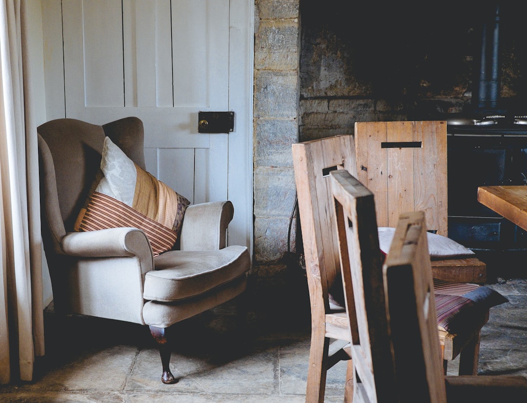 A photo of an armchair and dining table in the corner of a rustic English cottage interior with vintage wooden furniture, stone walls, warm light, and muted colours, shot on Fujifilm Provia film stock in the style of by an unknown artist. –ar 128:97