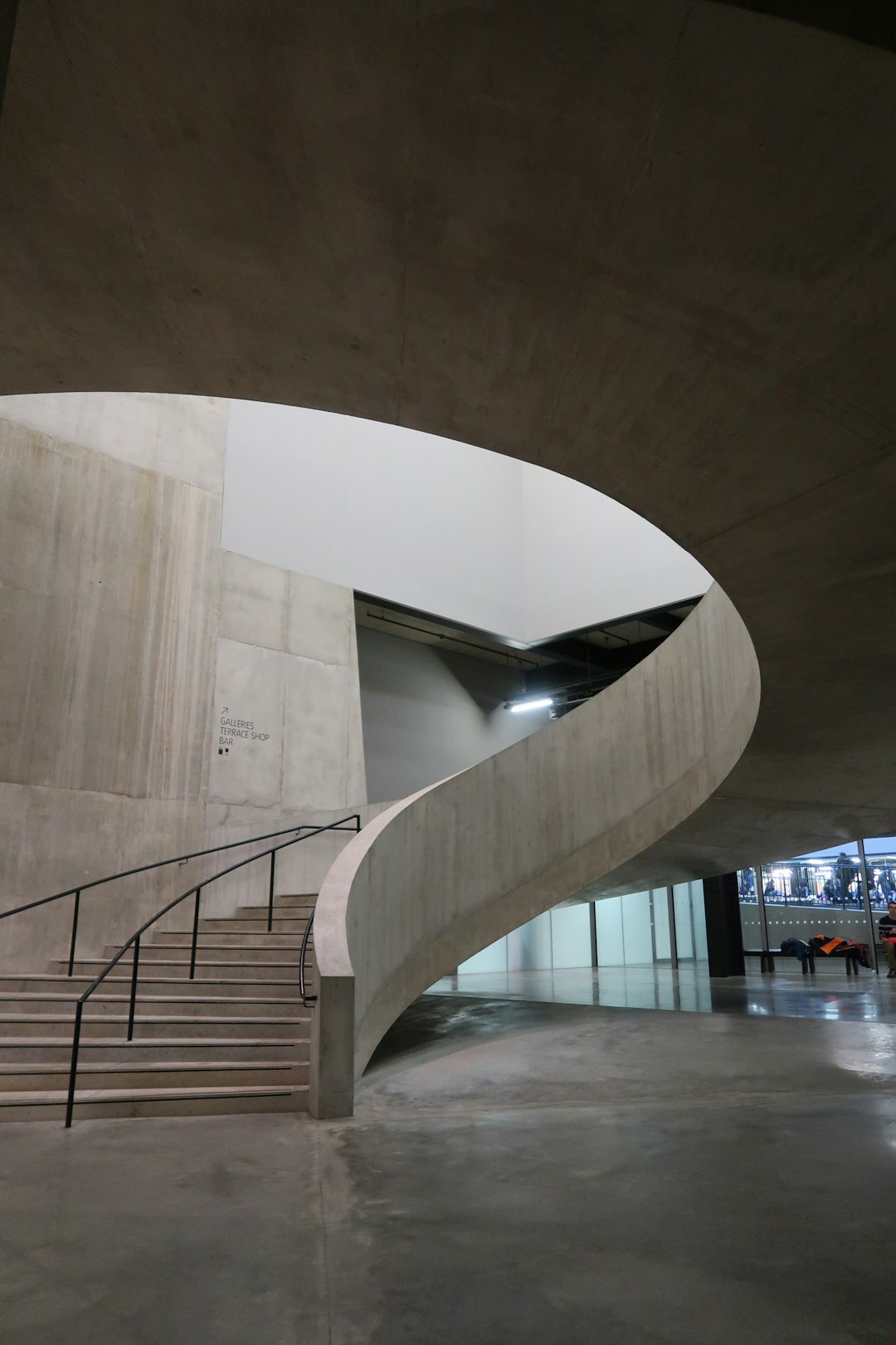 The interior of the new museum building features an undulating concrete wall with curved staircases leading to various areas, creating a strong sense of design and artistry. The overall space is modern in style, with white walls, grey floors, and dim lighting, giving it a unique ambiance. There is also some furniture inside that adds depth and character to its appearance.