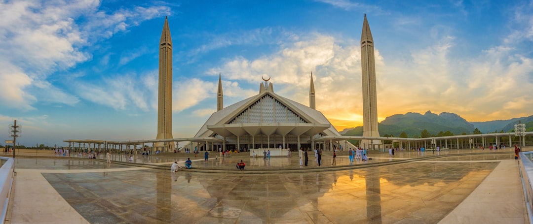 A panoramic view of the symmetrical landscape of an enchanted mosque and mountain, with golden hour light. The wide angle view has a clear sky and blue colored clouds. Crowds are walking around in front of the building in the style of [Feng Zhengjie](https://goo.gl/search?artist%20Feng%20Zhengjie). The photorealistic and super detailed scene has high resolution and HDR effects, as if shot on a Sony Alpha A7 III camera. –ar 128:53