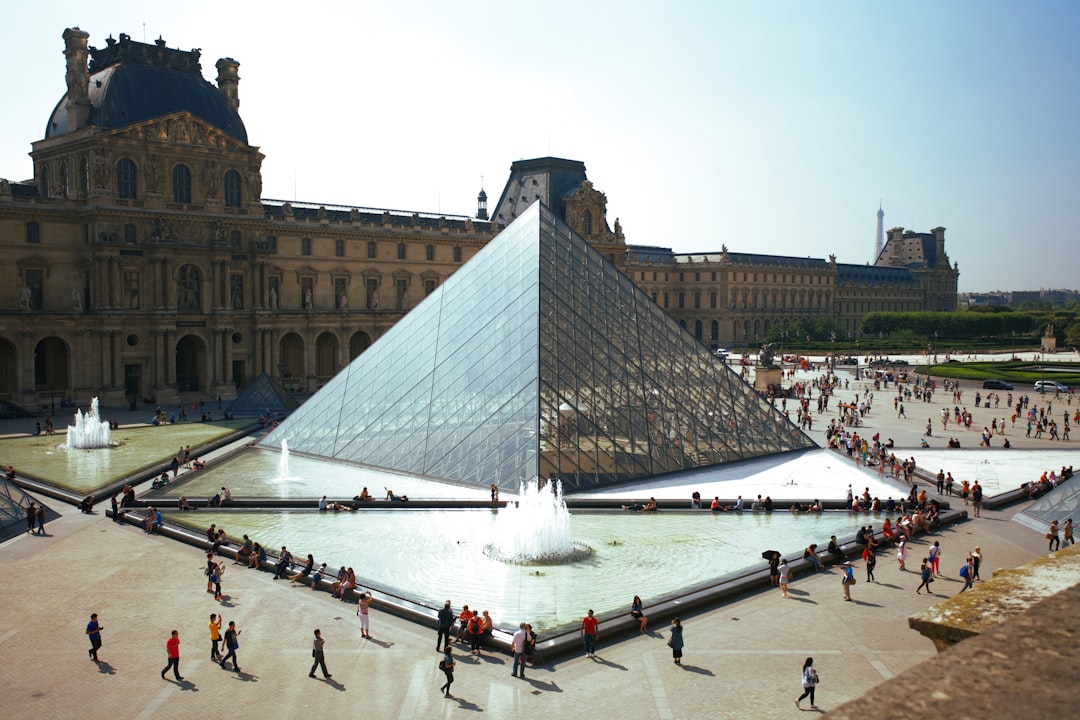 the Louvre Museum in Paris, with the large glass pyramid visible from outside. The square around it is crowded and full of tourists walking. In front of that there’s an empty pool filled with water. A fountain at one end sprays out into the air, creating a beautiful scene. This photo was taken by [Iwan Baan](https://goo.gl/search?artist%20Iwan%20Baan). –ar 128:85