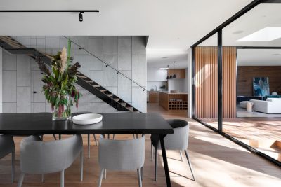 A photo of the dining room table in an open plan house with concrete walls, wooden floors and glass doors leading to another living space. The kitchen is visible through one wall and there's natural light streaming in from above. A staircase leads up to upper floor rooms. There are grey chairs around the black dinner table and vases full of fresh flowers on top of it. In front of that lies a white bowl filled with salad. Modern architecture photography in the style of an unnamed artist.