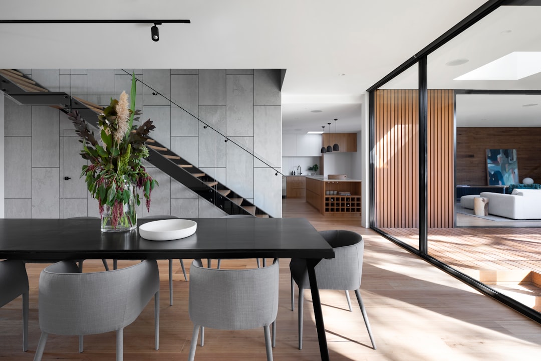 A photo of the dining room table in an open plan house with concrete walls, wooden floors and glass doors leading to another living space. The kitchen is visible through one wall and there’s natural light streaming in from above. A staircase leads up to upper floor rooms. There are grey chairs around the black dinner table and vases full of fresh flowers on top of it. In front of that lies a white bowl filled with salad. Modern architecture photography in the style of an unnamed artist.