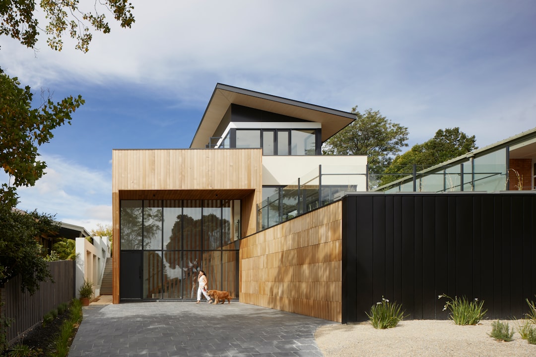 An architecturally designed home in Melbourne with timber cladding and black walls, glass windows and an entrance that leads to the garage where there is one dog running around inside. The house has two stories with flat roofs on top of it and concrete floor at ground level, and trees and plants surround its surroundings. A woman stands outside holding her small yellow puppy while another large brown Labrador runs through front door. Shot in the style of canon eos r5. –ar 128:85