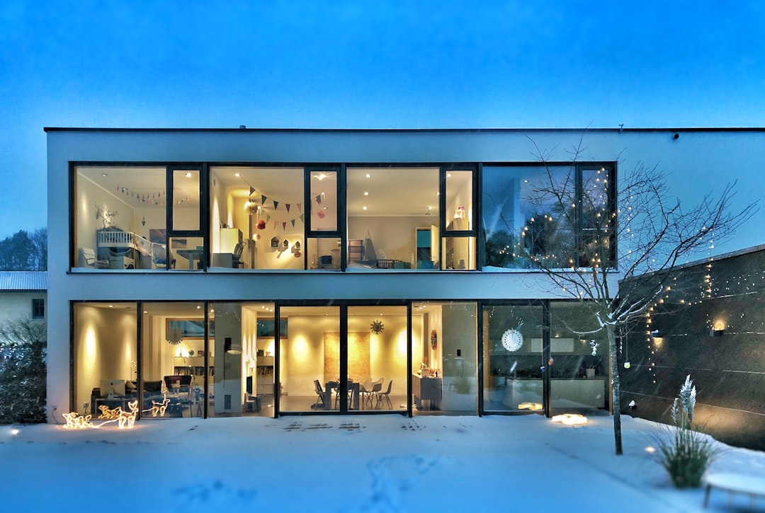 A modern house with large glass windows and white walls, illuminated by warm lights inside during winter in the early evening, showcasing an open living room on one side of two floors and a dining area on another side, surrounded by snow-covered gardens outside. The scene is captured from across the street at night, emphasizing the contrast between the cool blue sky and bright interior lighting. There is also some mist or fog adding to the atmosphere. The style of the scene is reminiscent of an artist known for paintings of modern homes illuminated from within against cool winter nights.