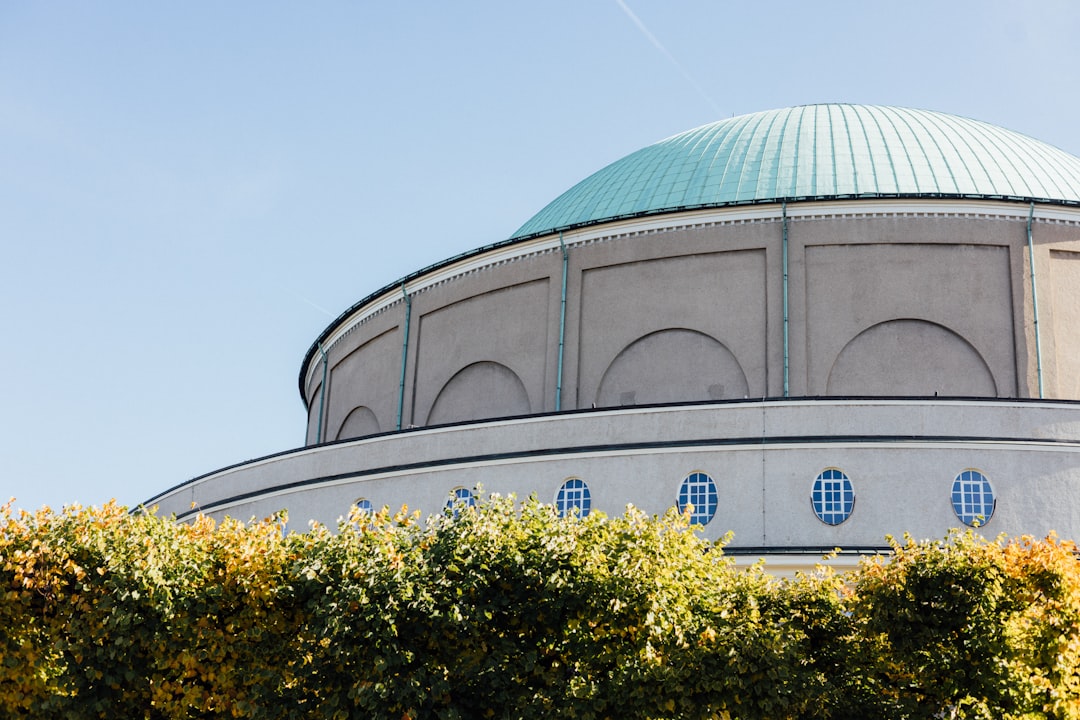 modern circular building with dome roof, light grey stone facade with blue and green accents, surrounded by bushes, blue sky in the background, architectural photography