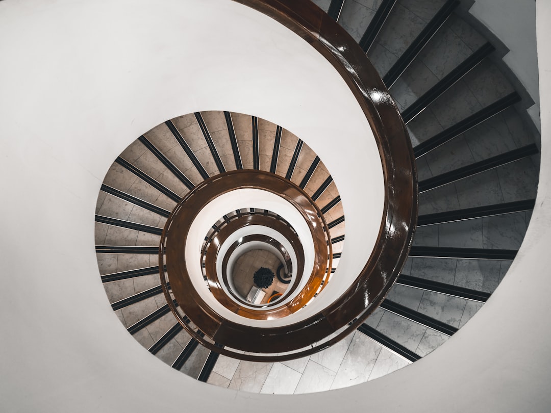 photo of a spiral staircase with white walls and a brown wood handrail, the view is from above looking down at a person walking up it, in the style of unsplash photography