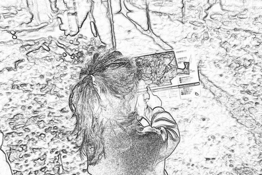 Black and white storyboard sketch of a young girl taking a selfie with a digital camera, showing her face in front view, wearing a ponytail hairstyle, with a cave wall made of stone in the background.