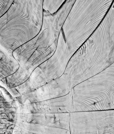 A closeup of an old, weathered wooden surface with visible grain patterns and swirling shadows, creating intricate textures in black and white. The wood has been aged by time, showcasing the beauty found within simplicity. This scene is captured from above, offering an overhead perspective that highlights the delicate interplay between light and shadow on each cross section of textured timber.