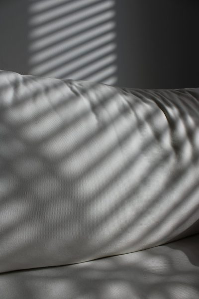 A white pillow with shadows of window blinds upon it, creating an abstract and minimalist composition. The focus is on the play between light and shadow in a simple yet elegant setting.