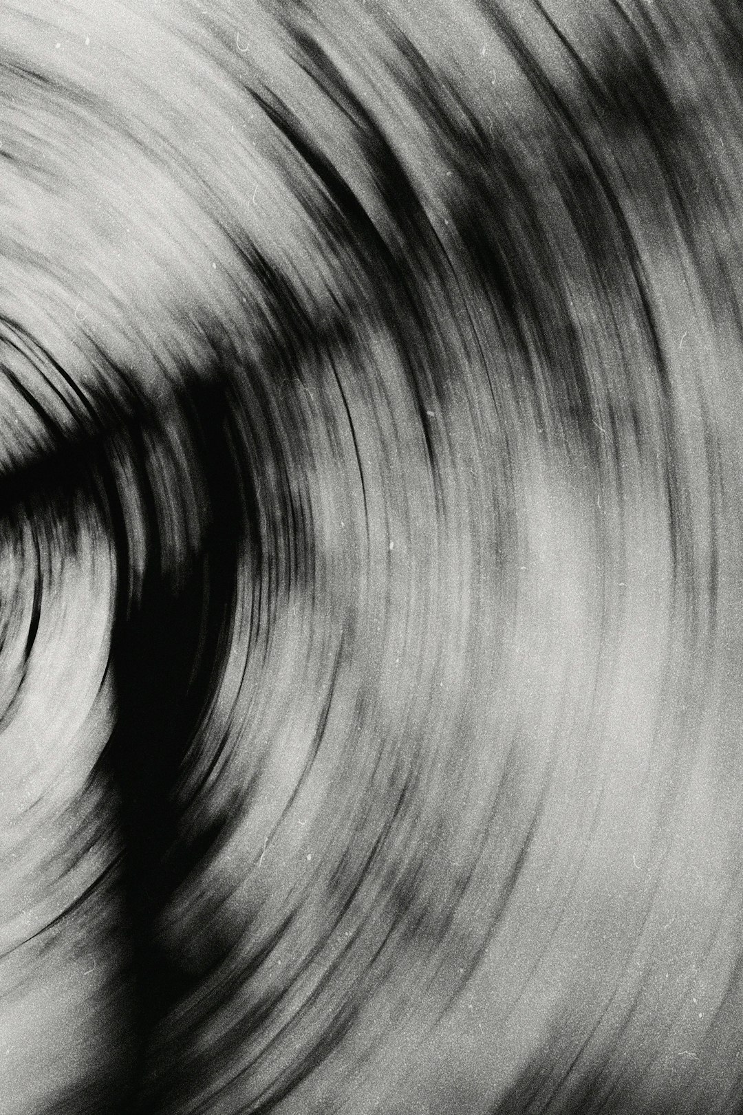 Abstract black and white photograph of an old vinyl record spinning, grainy texture, blurred motion, slow shutter speed, monochromatic tones with shades of gray and charcoal, focus on the vint circular patterns created by rotating grooves, creating visual effects reminiscent of wind and water ripples. The background is blurred to emphasize these intricate designs, evoking nostalgic memories or capturing the essence of music in mid tromp –ar 85:128