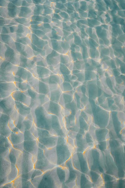 A top-down view of the clear, calm water in an outdoor swimming pool with ripples and reflections. The light reflects off the surface creating patterns that make it look like fine sand on a beach or a starry night sky. It is a beautiful scene of tranquility and beauty. The photo was taken from above looking down at the water. --ar 85:128