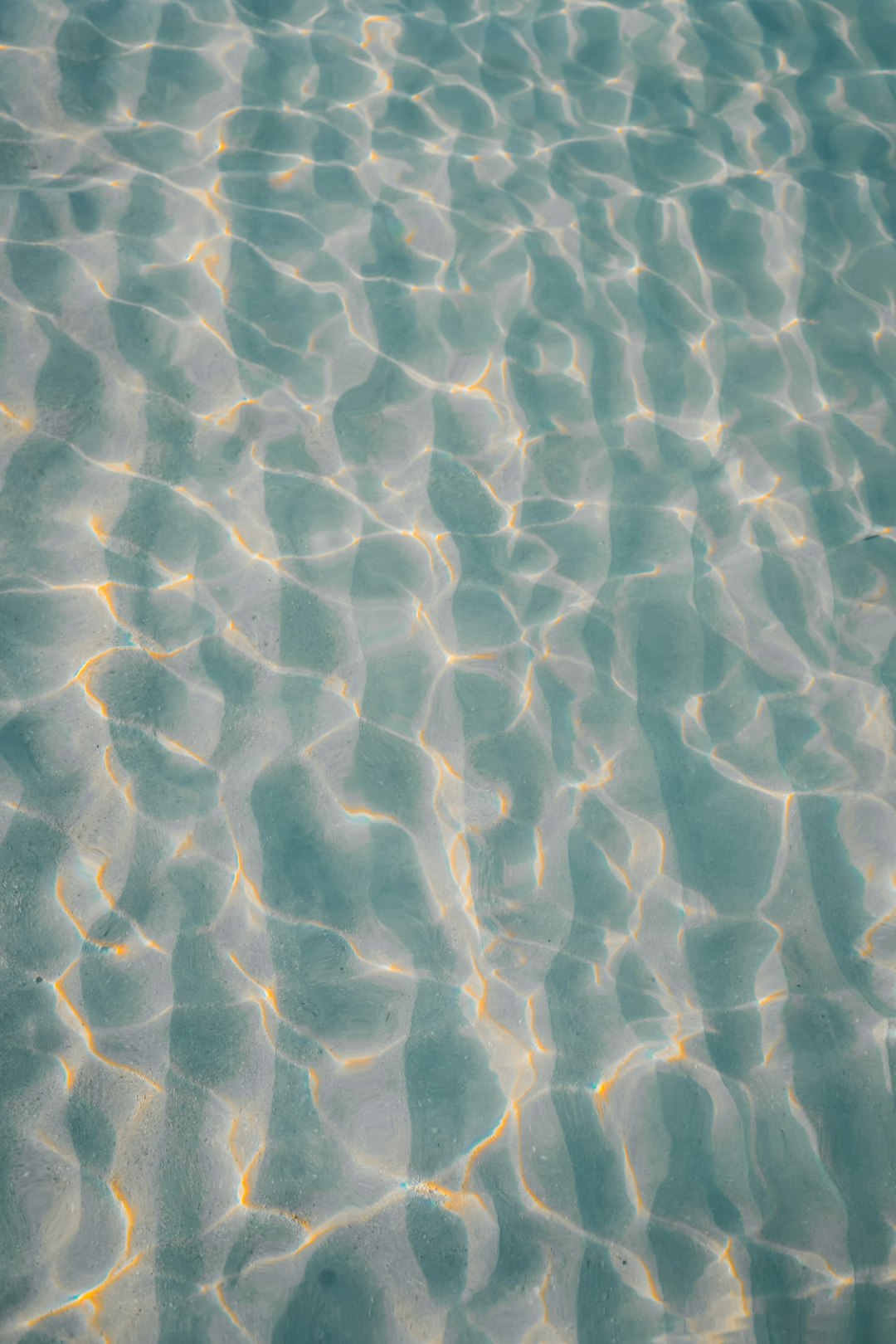 A top-down view of the clear, calm water in an outdoor swimming pool with ripples and reflections. The light reflects off the surface creating patterns that make it look like fine sand on a beach or a starry night sky. It is a beautiful scene of tranquility and beauty. The photo was taken from above looking down at the water. –ar 85:128