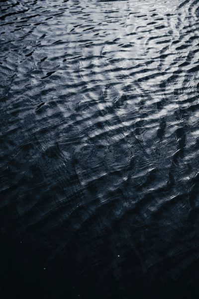 Dark water surface, ripples on the water's edge, closeup of ripples in dark blue tones, low saturation, low light reflection on wet ground, cool tone, top view angle, high contrast, mysterious atmosphere. --ar 85:128