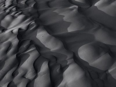 A highresolution black and white photograph of the texture details in sand dunes, captured from above with dramatic lighting that casts deep shadows on its rugged surface. The composition focuses on undulating patterns created by wind, showcasing intricate textures and shapes in every grain of dust. This photorealistic image highlights how the desert's terrain is not just flat but full of life, capturing both depth and beauty in the mesmerizing interplay between light and shadow. --ar 4:3