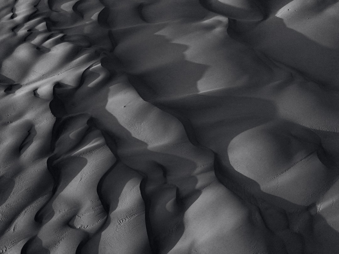 A highresolution black and white photograph of the texture details in sand dunes, captured from above with dramatic lighting that casts deep shadows on its rugged surface. The composition focuses on undulating patterns created by wind, showcasing intricate textures and shapes in every grain of dust. This photorealistic image highlights how the desert’s terrain is not just flat but full of life, capturing both depth and beauty in the mesmerizing interplay between light and shadow. –ar 4:3