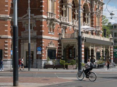 A woman is riding her bike in the city of Amsterdam. As she passes by an elegant hotel with ornate architecture and large windows, on one side stands out a street lamp. Next to it, there are also some people walking around and sitting at outdoor cafes at tables. The road has red brick pavement and white lines drawn along its edges. Above them you can see several signs indicating different streets or squares. In front of us are two black metal pillars that support an overhanging bridge. --ar 4:3