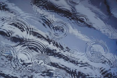 Raindrops creating circular ripples on the water surface, making abstract patterns and reflections in the sky. --ar 128:85