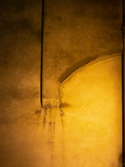 A closeup of the curved wall in an old, dimly lit room with yellowish lighting casting long shadows on its surface. The wall is painted beige and has subtle textures that give it depth. A small crack or smile mark can be seen at one corner where two walls meet, adding to the overall mysterious atmosphere. --ar 3:4