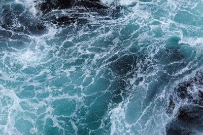 Closeup of the rough sea, aerial view, dark blue and turquoise colors, top down perspective, waves crashing against rocks and splashing water, dynamic movement, high resolution photography, hyperrealistic details, natural lighting, detailed texture with a focus on the ocean surface from a high angle camera position, in the style of naturalistic photography. --ar 128:85