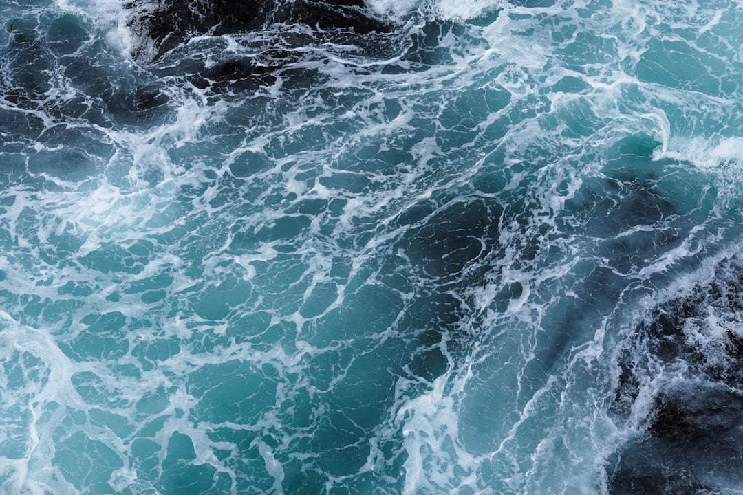 Closeup of the rough sea, aerial view, dark blue and turquoise colors, top down perspective, waves crashing against rocks and splashing water, dynamic movement, high resolution photography, hyperrealistic details, natural lighting, detailed texture with a focus on the ocean surface from a high angle camera position, in the style of naturalistic photography. –ar 128:85