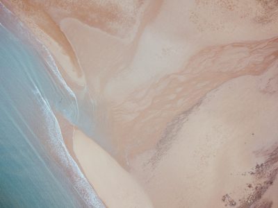 Aerial view of the sandy beach, with smooth lines and water ripples in shades of light brown, pink, bluegreen, and white, creating an abstract composition that evokes calmness and tranquility. The image captures natural beauty and serenity, adding to its overall aesthetic appeal. This aerial shot provides a unique perspective on the beach's landscape, emphasizing patterns created in the style of gentle waves and sand dunes. --ar 128:95