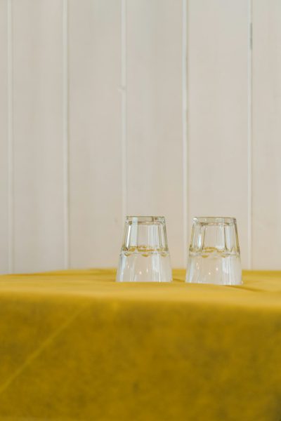 Two empty glasses on a yellow tablecloth in front of a white wall, minimalistic wedding venue photography in the style of minimalism. --ar 85:128