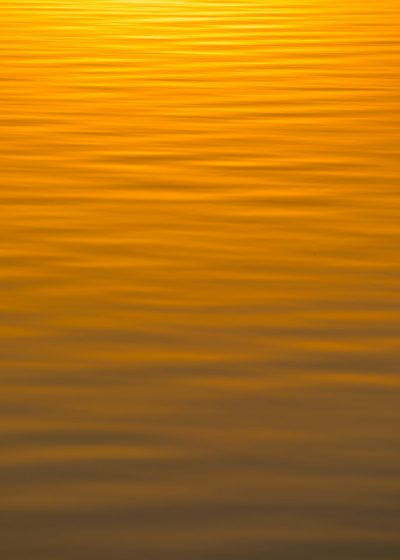 Golden sunset light reflecting on the water surface, creating ripples and golden stripes in an abstract background. The image has soft tones of orange to yellow with no visible objects or textures. It's a highresolution, highquality photo that captures the serene beauty of nature at dusk. This is suitable for artistic or design purposes, such as a wallpaper or banner. --ar 91:128